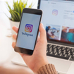 Closeup of woman holding smartphone logged into Instagram in front of laptop