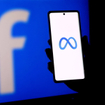 A person holding a smartphone displaying the meta logo in front of a facebook logo screen.