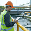 Environmental engineer in a reflective vest inspecting a wastewater treatment plant with a digital tablet.