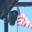 Flock camera monitoring a parking lot as an american flag waves in the background.