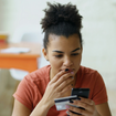 A young woman contemplates her phone with bank card in hand.