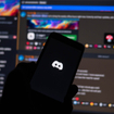 Discord logo on phone screen, with the desktop app opened on a monitor in the background.