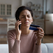 Frustrated woman sitting on couch at home trying to cancel subscription over the phone while holding a credit card