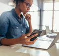 A young Black woman holds her smartphone in her right hand while looking at her laptop screen