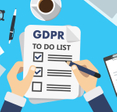 An illustration of a man&#x27;s hands holding a piece of paper with a GDPR to-do list on it. The background is a messy desktop with glasses, a cup of coffee, papers, and a laptop.