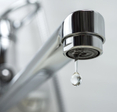 A close-up photo of a stainless steel kitchen sink faucet with a single drop of water leaking out of it.