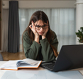 A young, dark-haired woman with glasses and a forest green sweatshirt holds her head in her hands and looks in frustration at her laptop.
