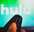 A close-up photo of a woman&#x27;s hand holding a remote and pointing it at her TV. On the screen is the Hulu logo.