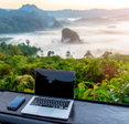 A laptop and cell phone sit on a table facing a gorgeous view of the sun rising over a jungle and mountains.