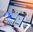 A young woman holds her smartphone that is showing payment success on screen in one hand and her credit card in another, while her laptop is on the table.