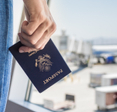 A close-up of a man&#x27;s hand holding a U.S. passport and standing in front of an airport window watching travelers board a white and blue airplane.