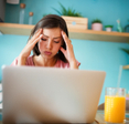 Woman with hands on her head looking frustrated while in front of laptop, glass of orange juice on the side and shelf on the back wall