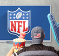 Person&#x27;s back as they wear American flag cap, popcorn and soda on one hand, and a giant foam hand in front of TV that shows NFL 
