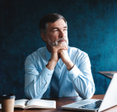 Focused older businessman deep in thought while sitting at a table in modern office with a laptop and a notebook in front of him.