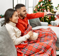 Young couple is sitting on the couch by their Christmas tree while the man is turning on the TV.