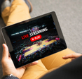 A person sitting on the ground holding a tablet in one hand and watching a livestream of a sports broadcast. The screen says &quot;Live Streaming&quot; and shows a basketball court.