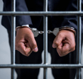 A close-up of a man&#x27;s hands in handcuffs as he&#x27;s behind a jail cell.