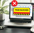 An open laptop on a work desk with the screen displaying the text &quot;page blocked&quot; over a restricted website
