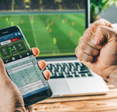A man holds a phone showing a sports score of 2 to 1 on a betting app. His right fist is clenched in victory. There&#x27;s a laptop screen in the background showing the soccer game being played.