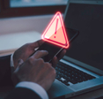 A man holding his smartphone in front of his laptop. There&#x27;s abstract warning above his phone.