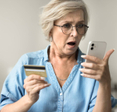 A shocked older woman is holding her smartphone in one hand and her credit card in another.