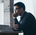 A man sits in front of his computer, resting his chin in his hand.
