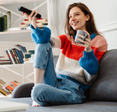 A woman sits on her couch, holding a mug and the remote.