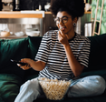 A woman sits on the couch, eating popcorn and watching TV.