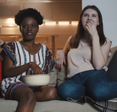 Happy young multiethnic women friends sit together at home watching movies smiling together with snacks slow motion.
