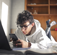 Teen boy laying on bed while scrolling on his smartphone and working on his laptop.