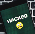 Closeup of a smartphone with a sad emoji and the word Hacked in all caps