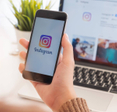Closeup of woman holding smartphone logged into Instagram in front of laptop