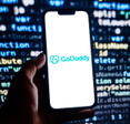 Closeup of smartphone displaying GoDaddy logo with code on a screen in background