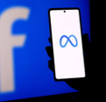 A person holding a smartphone displaying the meta logo in front of a facebook logo screen.