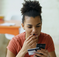 A young woman contemplates her phone with bank card in hand.