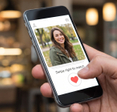 Close-up of a hand holding a smartphone displaying a dating app profile with a smiling woman and a 'Swipe right to match' button in a cafe setting.