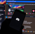Discord logo on phone screen, with the desktop app opened on a monitor in the background.