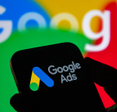 Closeup of a hand holding a smartphone displaying the Google ads logo with the Google logo in the background.