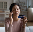 Frustrated woman sitting on couch at home trying to cancel subscription over the phone while holding a credit card