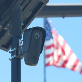 Flock camera monitoring a parking lot as an american flag waves in the background.