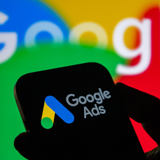 Closeup of a hand holding a smartphone displaying the Google ads logo with the Google logo in the background.