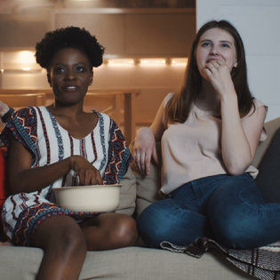 Happy young multiethnic women friends sit together at home watching movies smiling together with snacks slow motion.