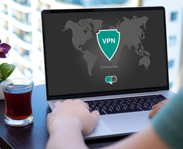 A person types on a laptop computer next to a glass of tea. The laptop screen shows a shield icon that says "VPN." The screen also shows a switch that shows the VPN is turned on.