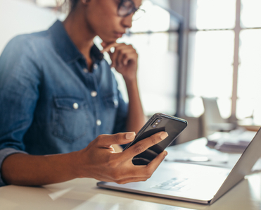A young Black woman holds her smartphone in her right hand while looking at her laptop screen