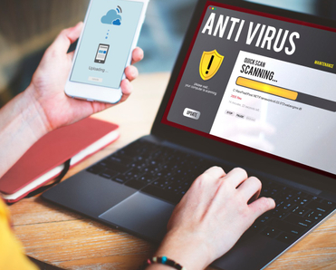 A woman&#x27;s hands hold her cell phone and type on her laptop while an antivirus software scan is on the screen
