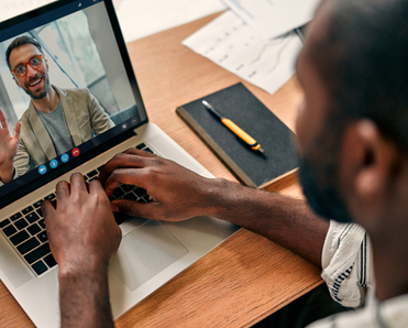 A rear-view of a Black man video conferencing with his coworker on Zoom. The desk is covered with paperwork, and the coworker on the screen is waving and smiling.