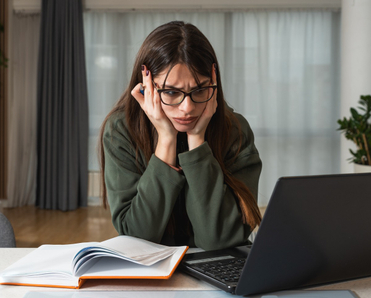A young, dark-haired woman with glasses and a forest green sweatshirt holds her head in her hands and looks in frustration at her laptop.