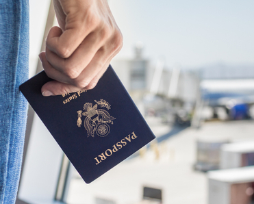 A close-up of a man&#x27;s hand holding a U.S. passport and standing in front of an airport window watching travelers board a white and blue airplane.