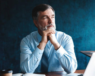 Focused older businessman deep in thought while sitting at a table in modern office with a laptop and a notebook in front of him.