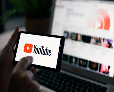 A smartphone being held with two hands with a laptop screen in the background. Both screens are showing YouTube content.